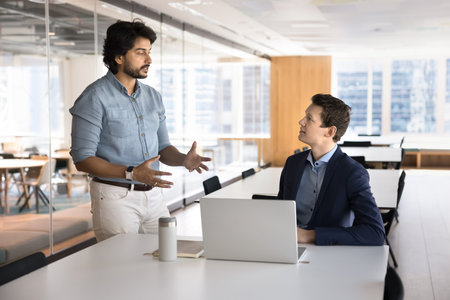 Knowledge sharing. Confident mature Indian man mentor team leader explain work details to millennial employee trainee teach male intern sitting by notebook guide young professional to career successの写真素材