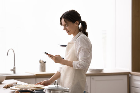 Digital help for home baking. Smiling young woman housewife in kitchen apron stand by countertop...