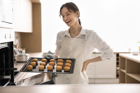 Home baking success. Proud smiling female in apron holding tray with fresh golden muffins standing...