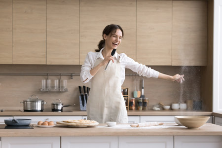 Home cooking karaoke. Overjoyed energetic millennial housewife enjoy carefree moment baking pizza pie turn spatula into microphone to sing song have fun snap fingers tossing flour dust in playful moodの写真素材