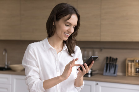 Digital connection. Portrait pretty smiling young girl stand at modern home kitchen interior type message on mobile phone enjoy pleasant online conversation in text chat stay connected browse websitesの写真素材