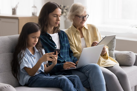Family screen dependence. Three female generations relatives older granny adult mom young daughter kid sit in line on cozy couch use different electronic devices networking at leisure time on weekendの写真素材