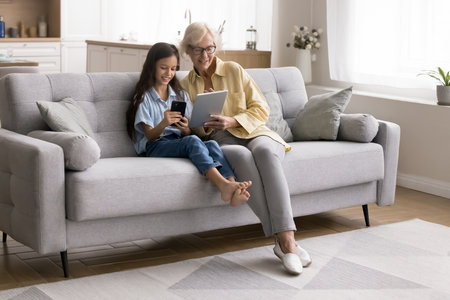Time for tech. Older adult grandmother little girl grandchild sit on comfy couch using diverse electronic devices exchange information via wireless internet play online education game on two gadgetsの写真素材
