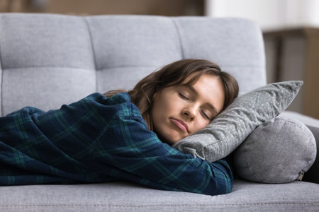 Sweet serene dreams. Tired millennial woman lie on stomach on soft comfy couch with eyes closed hugging pillow relax at daytime to recharge energy during peaceful nap enjoy restful sleep at cozy homeの写真素材