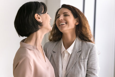 Affectionate moment. Two diverse age females middle aged woman mother teacher young daughter student stand indoors together hug laugh share happiness love warmth devotion through emotional eye contactの写真素材