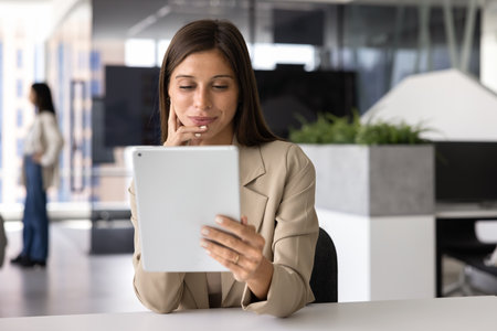Positive young Latin businesswoman watching online content on social media, using tablet computer at workplace, reading text on Internet, touching chin, thinking, sitting at office tableの写真素材