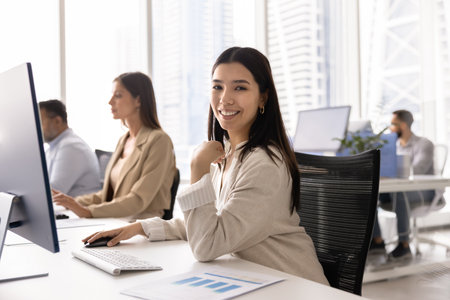 Happy Asian marketing agency analyst working at computer, looking at camera, smiling for portrait, sitting at workplace with large monitor, paper reports and diverse colleagues in backgroundの写真素材