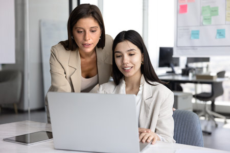 Young female business manager, mentor, consultant helping young Asian employee with online project, supervising work of staff, standing at laptop of coworker, looking at screen, explaining taskの写真素材
