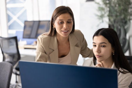 Two positive diverse female coworkers looking at computer monitor together, meeting at workplace for teamwork. Business expert woman giving consultation to young professional at workplaceの写真素材