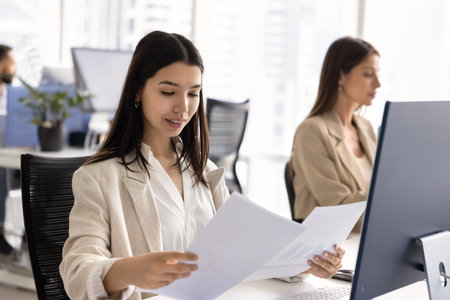 Busy young marketing analyst woman reviewing paper reports at workplace, analyzing research results, working on analytic data in open office workspace. Professional checking financial documentsの写真素材