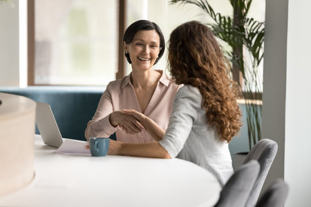 Smiling mid aged female client shake hands with young insurance agent finance advisor make agreement grateful for professional consultation. Older colleague appreciate younger teammate for mentoringの写真素材