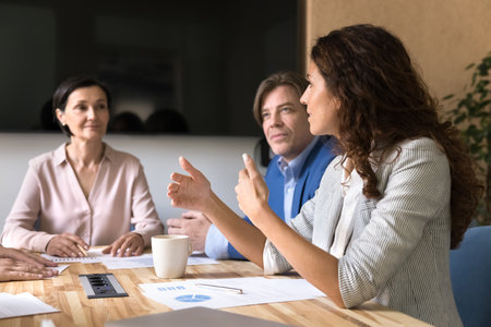 Presenting fresh strategy. Smart young businesswoman project manager executive talk to diverse mature colleagues at conference table express idea explain proposal share opinion based on statistic dataの写真素材