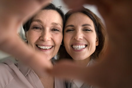 Celebrating family love. Affectionate portrait cheerful excited faces of joyful senior mother happy grownup daughter cuddle close smiling at camera through tender heart shape formed by joint fingersの写真素材
