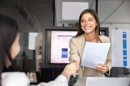 Happy young Hispanic business woman shaking hands with female coworker, smiling, holding paper document. Positive project manager giving handshake to partner after presentationの写真素材