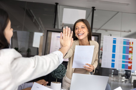 Positive young Latin business leader woman giving high five hand to female colleague, standing and bending over meeting table, holding paper document, smiling, laughing, thanking for good jobの写真素材
