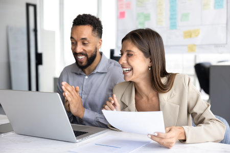 Two cheerful diverse office colleagues and friends having fun at workplace, discussing creative ideas for Internet sales growth, sitting at laptop, laughing, enjoying teamwork, corporate friendshipの写真素材