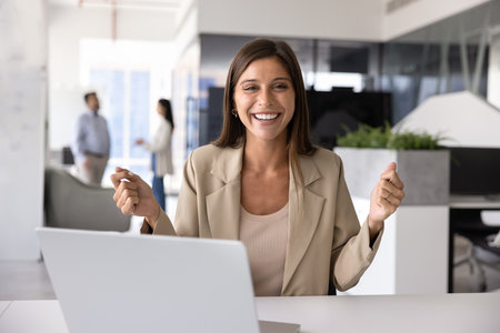 Cheerful young company employee woman celebrating business success at workplace with laptop, making hands winning gesture, looking at camera, laughing, getting good amazing newsの写真素材
