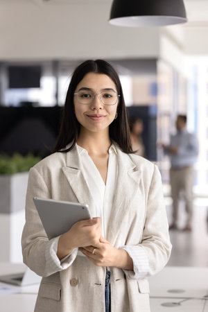 Happy successful smart young Asian professional woman in stylish eyeglasses holding tablet pc, posing for vertical business portrait, looking at camera, smiling. Vertical shotの写真素材