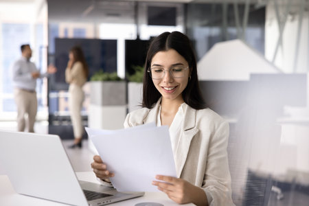 Happy young Asian financial professional woman in glasses reviewing paper reports at work table, smiling, analyzing growth, income, doing paperwork, reading analytic data, bill, invoiceの写真素材