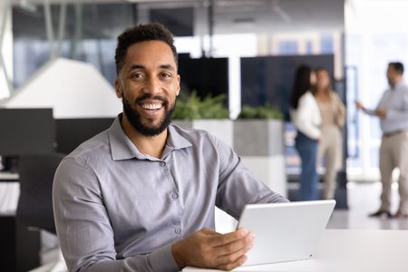 Positive African American manager posing at office workplace, holding tablet computer, looking at camera with toothy smile. Happy successful young business professional portraitの写真素材