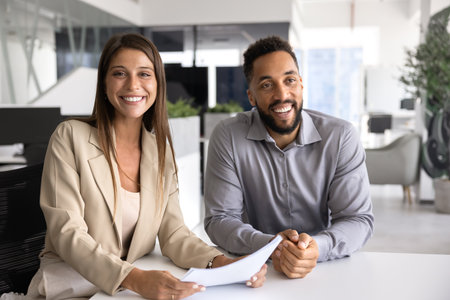 Two happy multiethnic business partners posing for portrait at office table, looking at camera and...