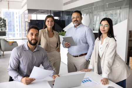 Diverse business team of four different aged professionals posing in office, standing at workplace table of one colleague, looking at camera, smiling. Grup portrait of company staffの写真素材