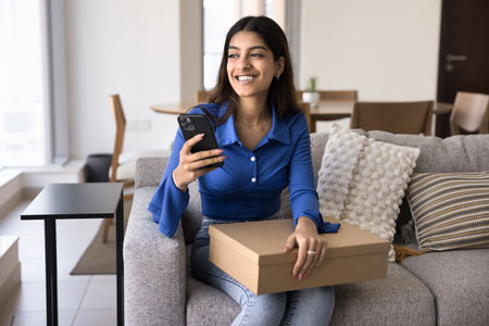 Cheerful young Indian customer woman holding received parcel, cardboard box at home, using mobile phone, making order, browsing online service, looking away, smiling, laughingの写真素材