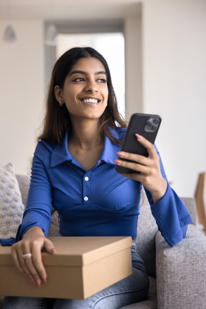 Positive young Indian woman using logistic application on smartphone, ordering delivery transportation service for sending parcel, holding postal cardboard box, looking away, smilingの写真素材