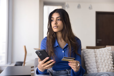 Serious young Indian customer woman shopping on ecommerce platform at home, making online payment by credit card, using bank application on mobile phone for transactionの写真素材