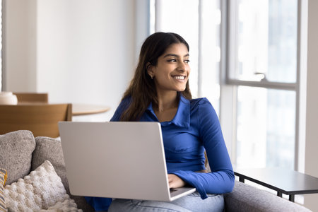 Positive young Indian freelance woman enjoying remote work from home, resting on soft couch, typing on laptop, looking at window away, smiling, thinking on creative solutions for projectの写真素材