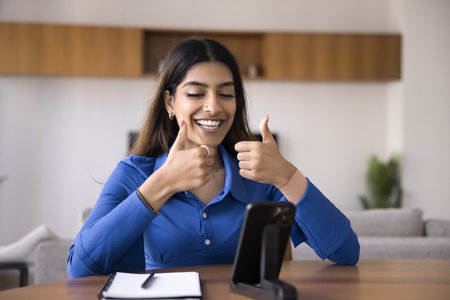 Cheerful beautiful young Indian woman showing double thumbs up at mobile phone, talking on online video conference call, smiling, using cellphone on holder for Internet communicationの写真素材