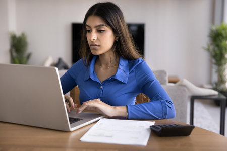 Focused young Indian freelance bookkeeper woman working on difficult accounting task, typing on laptop, looking at screen carefully, sitting at home table with calculator ad documentsの写真素材