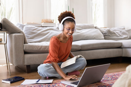Happy young adult African American student girl taking training course at home, watching learning webinar on laptop, using wireless headphones and computer, sitting on heating floorの写真素材