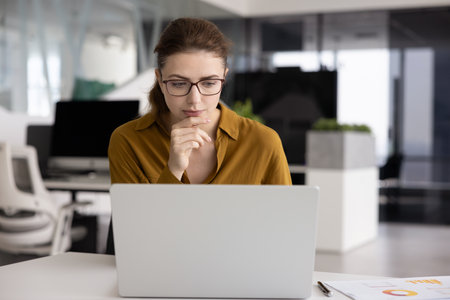 Serious concentrated young business professional woman working on online project at laptop, touching chin, thinking on creative solution, challenging task, Internet marketing strategyの写真素材