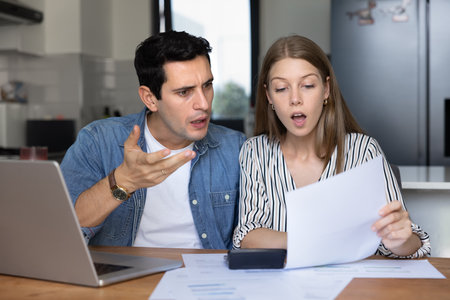 Young married couple reacting to financial document with shock and frustration, reviewing bank...