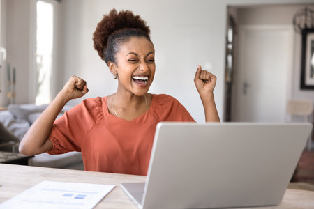 Excited joyful young African American business project manager making yes hand gesture at laptop, celebrating victory, professional triumph, successful startup, laughing, shouting for joyの写真素材