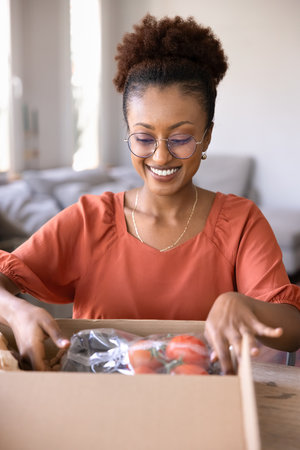 Happy young African American supermarket customer woman unpacking box with fresh vegetables, organic food, getting parcel from courier service, farm market. Vertical shotの写真素材