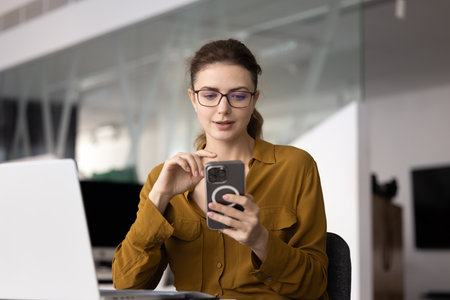 Positive young professional girl engaged in online communication, typing on smartphone at workplace,...