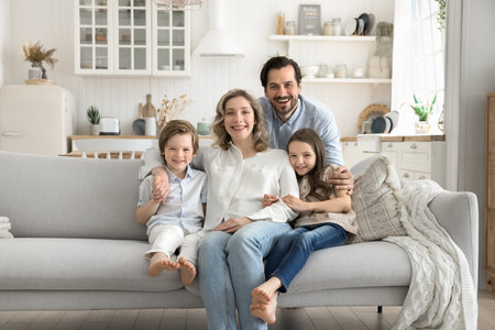 Cozy portrait smiling mom posing on couch in warm hugs of little daughter son loving dad. Friendly family with diverse kids relax on sofa look at cameraの写真素材