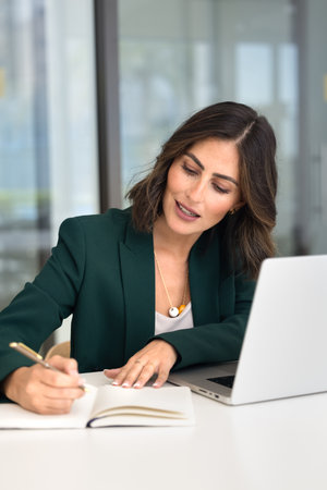 Busy young company CEO woman writing in notebook at workplace, planning task, takin notes, watching online training course on laptop pc, talking on video conference callの写真素材