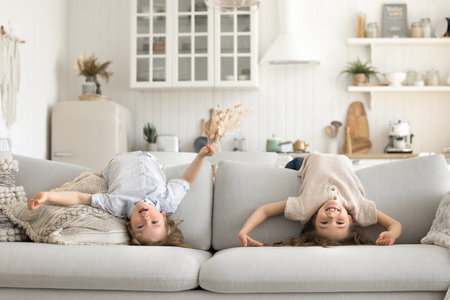 Upside down fun. Cute diverse little siblings messing hanging over back of large modern sofa in sunny home kitchen enjoy playing active game on durable comfy furniture designed for family lifeの写真素材