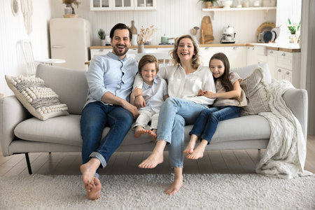 Sweet family home. Relaxed cheerful parents with 2 adorable kids little daughter son hug on couch look at camera with laughter. Portrait happy proud new homeowners tenants enjoy quality time togetherの写真素材