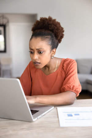 Shocked worried young African project manager working from home, typing on laptop, getting problems, bad news, finding mistake, failure, financial loss, staring at compute monitor. Vertical shotの写真素材