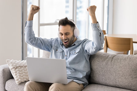 Happy young man in wireless headphones sits on sofa with laptop, raising arms in triumph and...