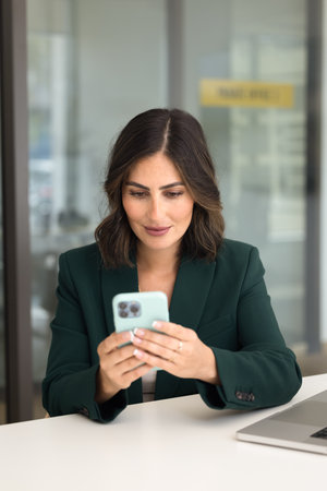 Positive beautiful young Hispanic business professional woman engaged in online communication typing on smartphone at office table, using agenda application for planning tasks. Vertical shotの写真素材