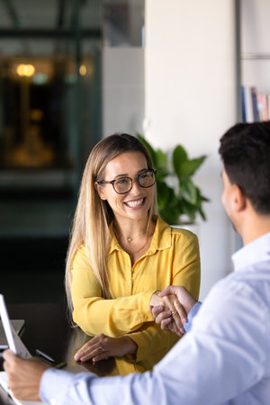 Cheerful young business professional woman in glasses getting hired, promoted, shaking hands with male colleague, boss, employer, thanking expert, consultant for help. Vertical shotの写真素材