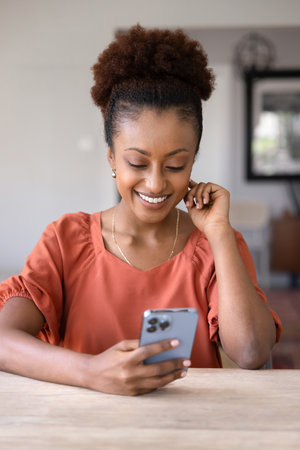 Happy young Black mobile phone user woman browsing Internet, social media on mobile phone, reading message, smiling, sitting at table, using smart app, web service. Vertical shotの写真素材