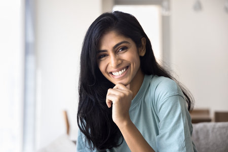 Head shot beautiful Indian woman posing for camera seated alone on cozy sofa at home, smiling, feel happy, enjoy carefree pastime at home at own or rented apartment. Tenancy, natural beauty, emotionsの写真素材