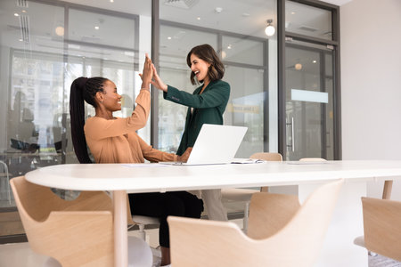 Two excited joyful diverse female colleagues celebrating teamwork success, making high five gesture at workplace with laptop, clapping hands, laughing, feeling cheerful after completing taskの写真素材