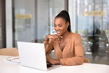 Happy young Black businesswoman typing on laptop at office table, touching chin, thinking on task, communication, using Internet technology for effective work on online projectの写真素材